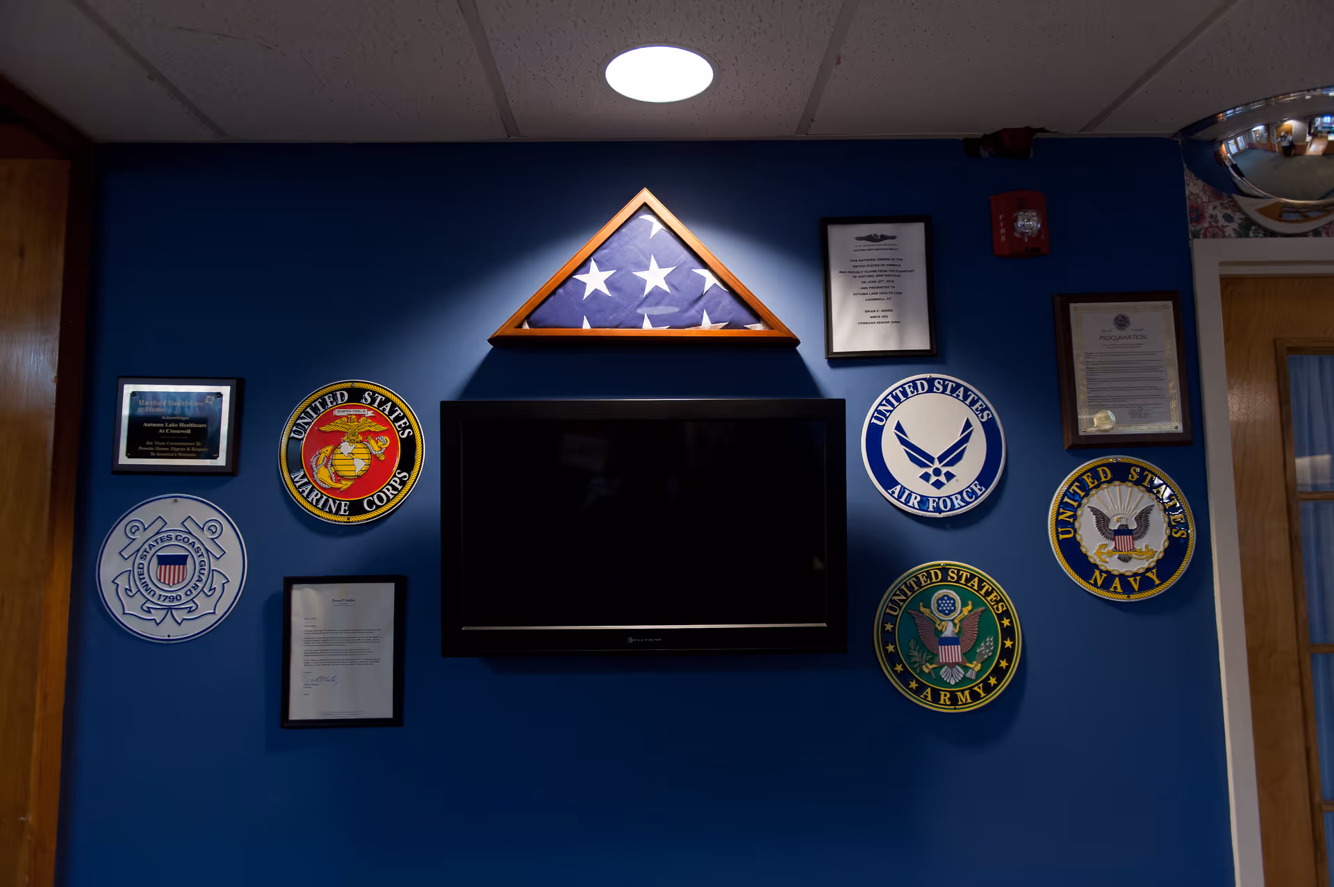 A blue wall displaying a folded American flag in a triangular wooden frame above a flat-screen TV. Surrounding the TV are plaques and emblems representing various United States military branches including the Marine Corps, Air Force, Coast Guard, Army, and Navy. There are also framed certificates and a fire alarm on the wall.