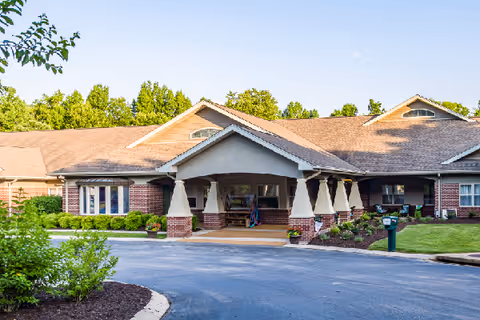 Front exterior view of a single-story senior living facility building with a covered entrance supported by columns, surrounded by landscaped greenery and a paved driveway.