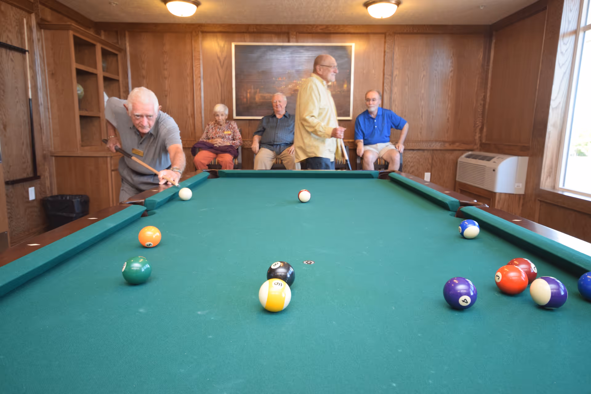 A group of elderly people in a wood-paneled room with a pool table. One man is aiming to take a shot with a pool cue, while three others sit on chairs against the wall and another man stands nearby holding a cue stick. The room has a large window and an air conditioning unit.