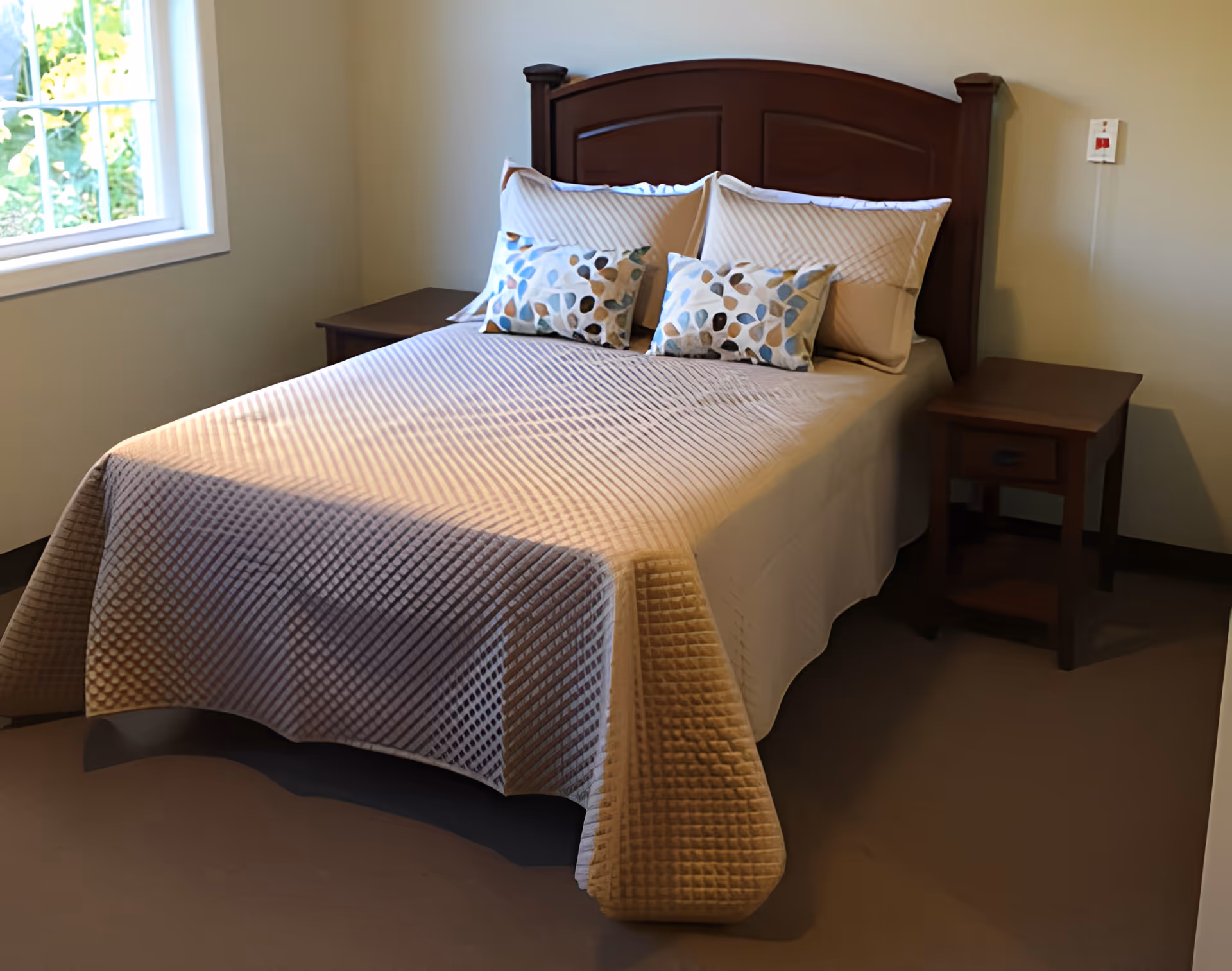 A neatly made bed with a dark wooden headboard, beige quilted bedspread, and four pillows including two decorative ones with a leaf pattern. The bed is flanked by two matching wooden nightstands. There is a window on the left side letting in natural light, and the walls are painted a light color.