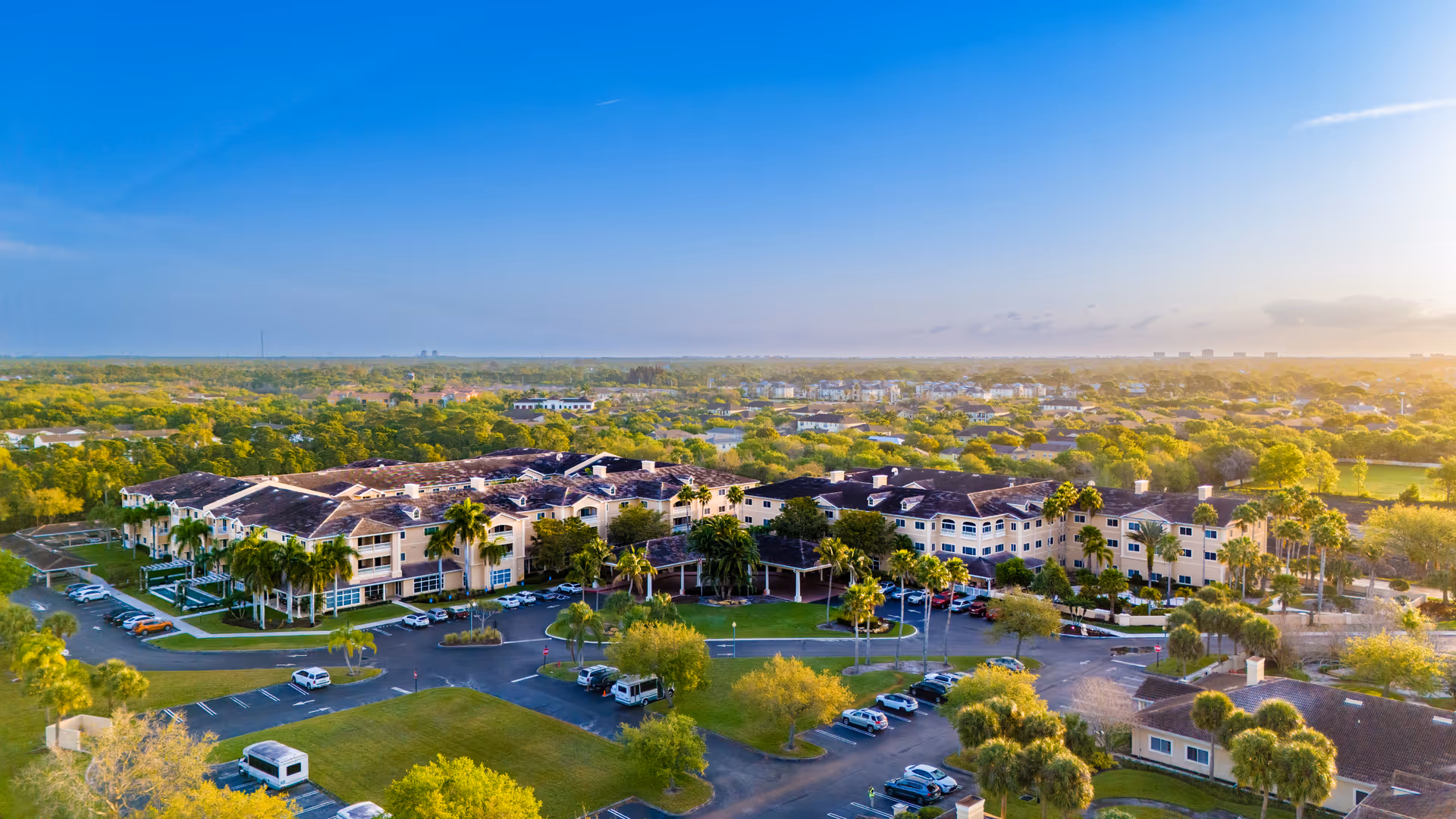 Aerial view of a large multi-wing senior living complex surrounded by trees, lawns, and parked cars under a clear blue sky.