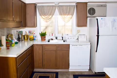 Small kitchen with wooden cabinets, a sink under a curtained window, white dishwasher and refrigerator, and various items on the countertops.