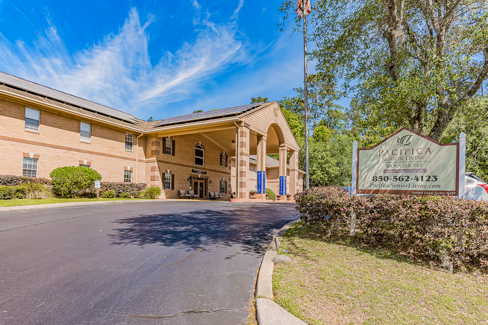 Exterior view of a senior living facility building with a covered entrance supported by columns. There is a sign in the foreground that reads 'Pacifica Senior Living Woodmont' with contact information and website. The building is surrounded by greenery and trees under a blue sky with some clouds.