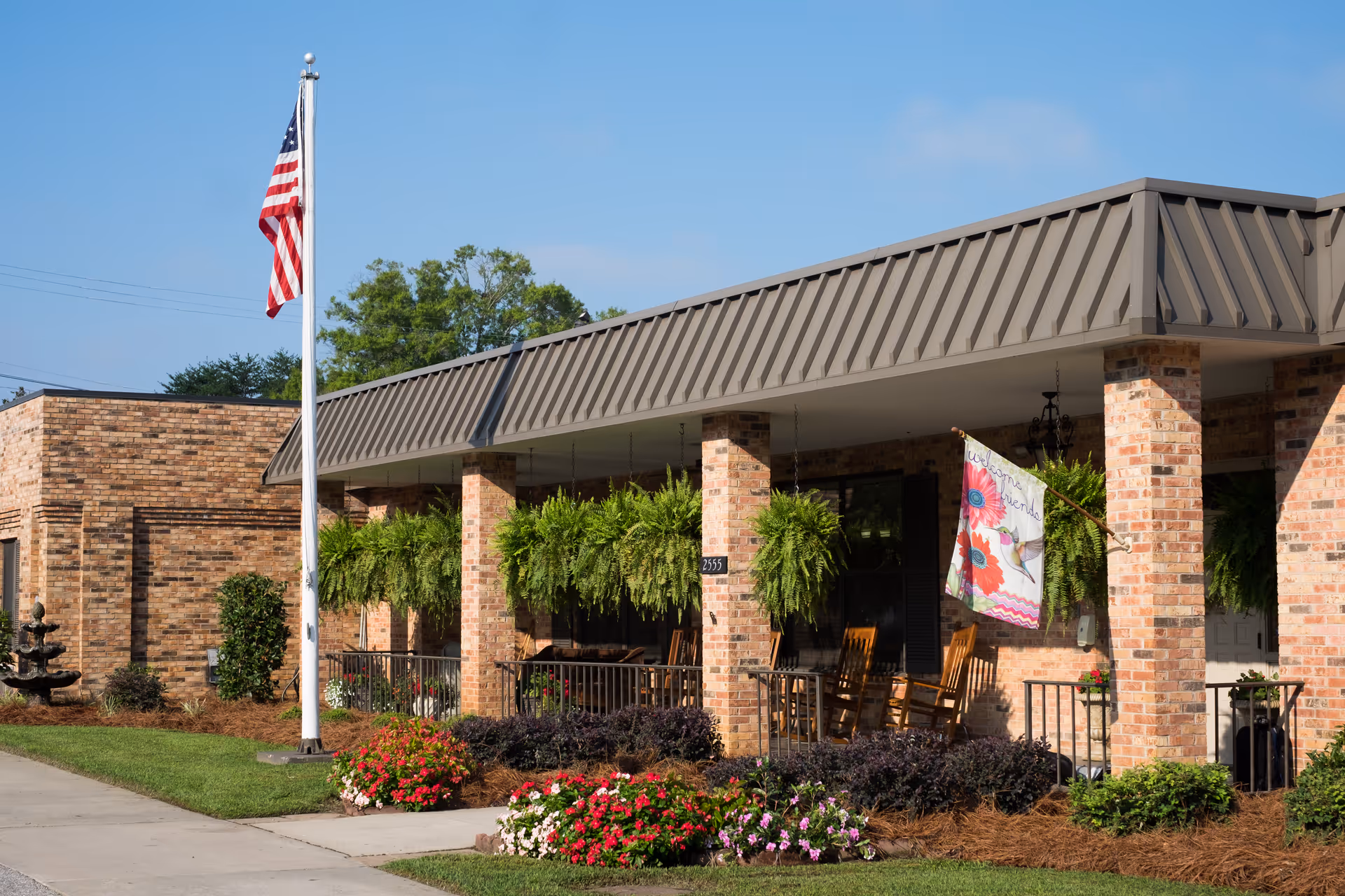 Exterior view of White Oak Manor Newberry showing a brick building with a covered porch. The porch has hanging green plants, rocking chairs, and a colorful welcome flag. An American flag is flying on a flagpole in front of the building, with well-maintained flower beds and shrubs along the walkway.