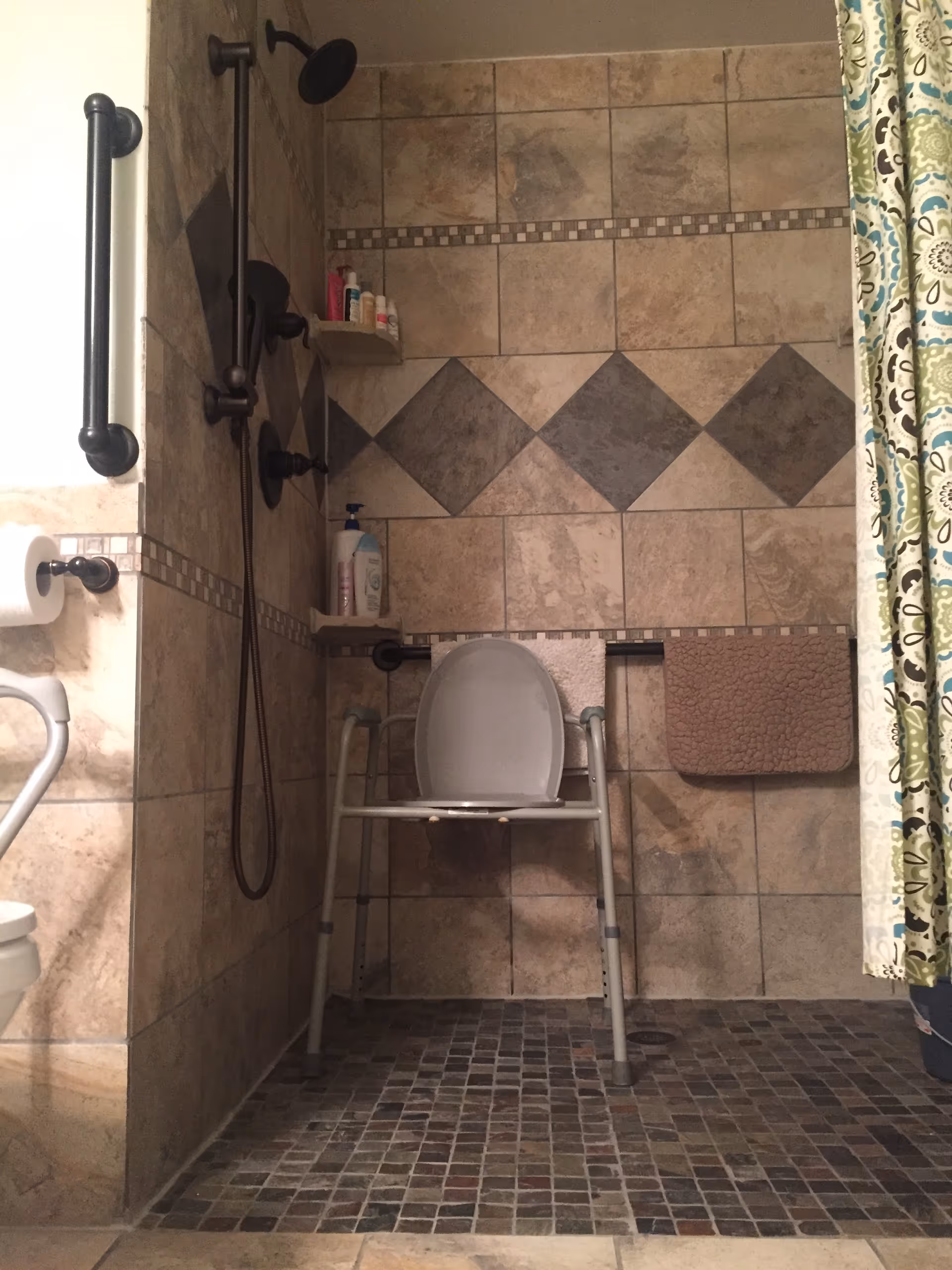 A tiled bathroom shower area with a shower chair, grab bars, a handheld showerhead, and shelves holding toiletries. There is a towel rack with two towels and a patterned shower curtain on the right side.