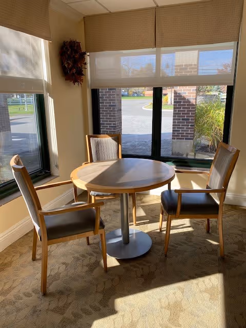 A small round wooden table with a metal base surrounded by three wooden chairs with cushioned seats and backs, placed near large windows with beige roller shades. Sunlight streams through the windows, casting shadows on the carpeted floor. Outside the windows, a brick column and some greenery are visible.
