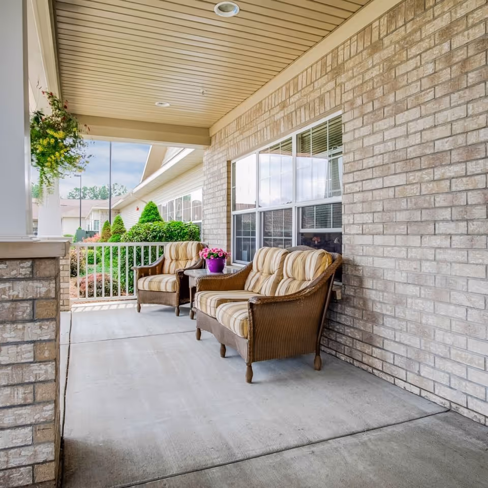 Covered front porch with two cushioned wicker chairs, a small table holding a purple potted plant, and a brick exterior wall.