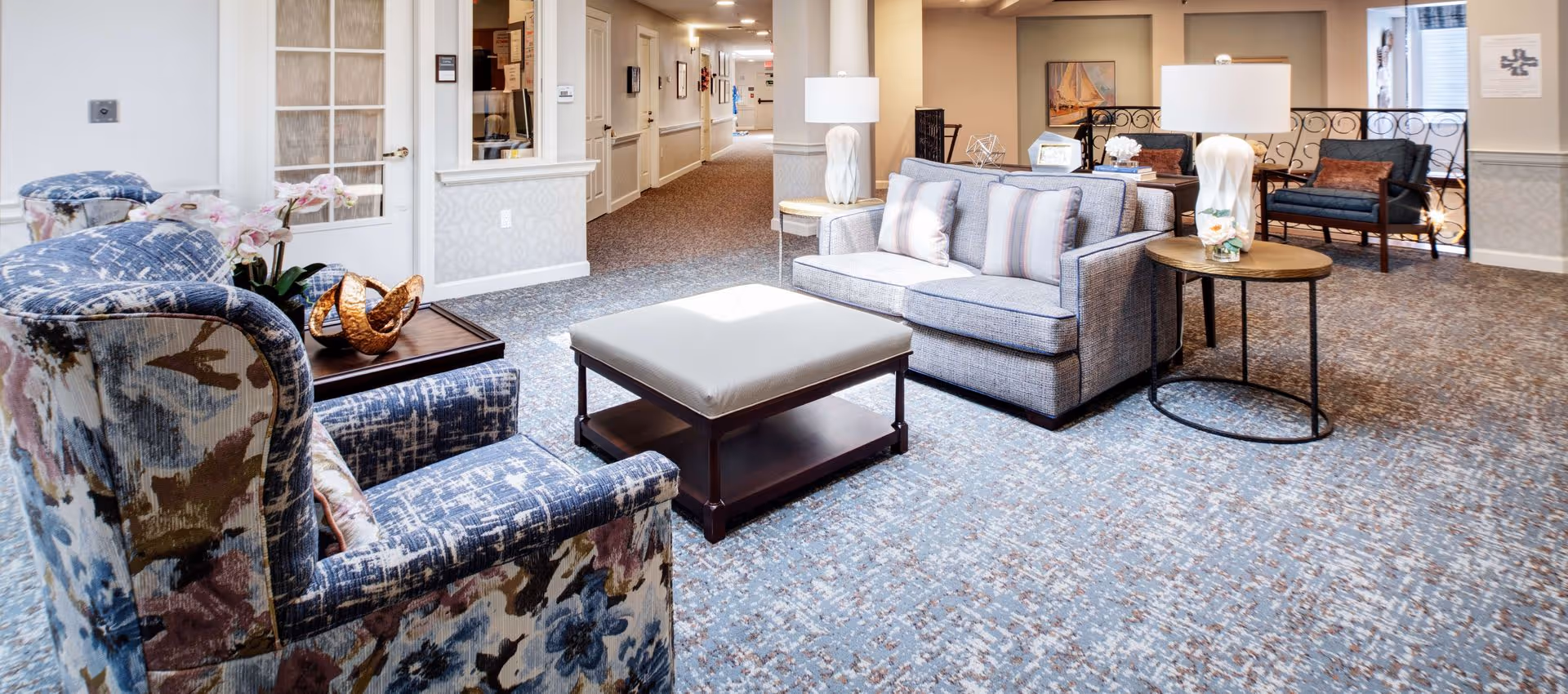 Bright, carpeted common lounge with upholstered armchairs, a sofa, ottoman and side tables in a senior living facility corridor.