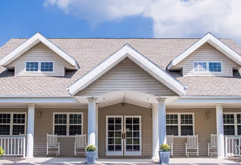 Front exterior view of a beige senior living facility building with a peaked roof, white columns, rocking chairs on the porch, and two potted plants near the entrance under a blue sky with some clouds.