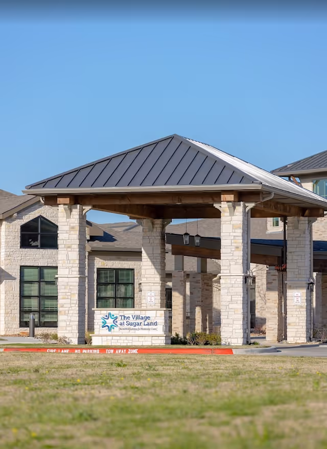 Exterior view of The Village at Sugar Land facility showing a covered entrance with stone pillars and a metal roof, with the facility sign displayed in front.