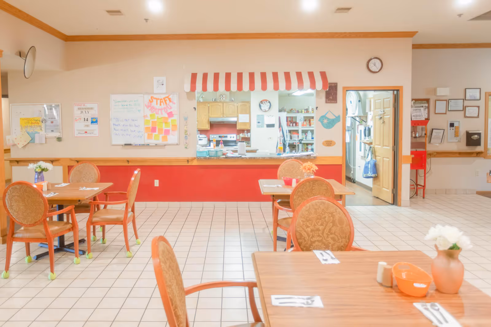 Bright dining room with tables and chairs facing a serving counter topped by a red-and-white striped awning.