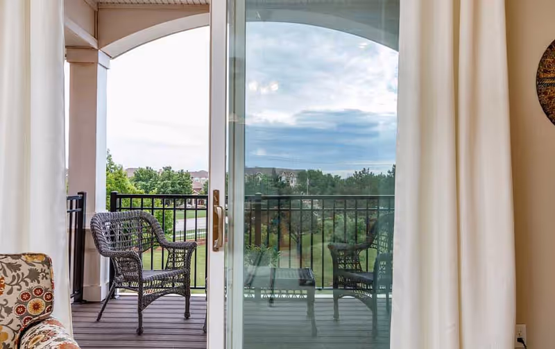 Sliding glass door opening onto a balcony with wicker chairs overlooking a grassy landscape and distant buildings.