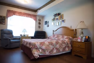A cozy bedroom in a senior living facility featuring a bed with a floral quilt, two recliner chairs near a window with pink curtains, a wooden nightstand with a lamp and decorative items, and framed pictures on the wall above the bed.