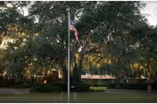 An American flag on a flagpole stands in front of large live oak trees and a low building façade.