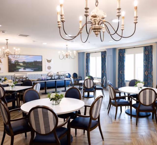 A bright dining room with round tables and cushioned chairs arranged neatly. Each table has a small floral centerpiece. The room features elegant chandeliers hanging from the ceiling, large windows with patterned blue curtains, and a buffet area with covered serving dishes along the back wall. The floor is wooden with a herringbone pattern.