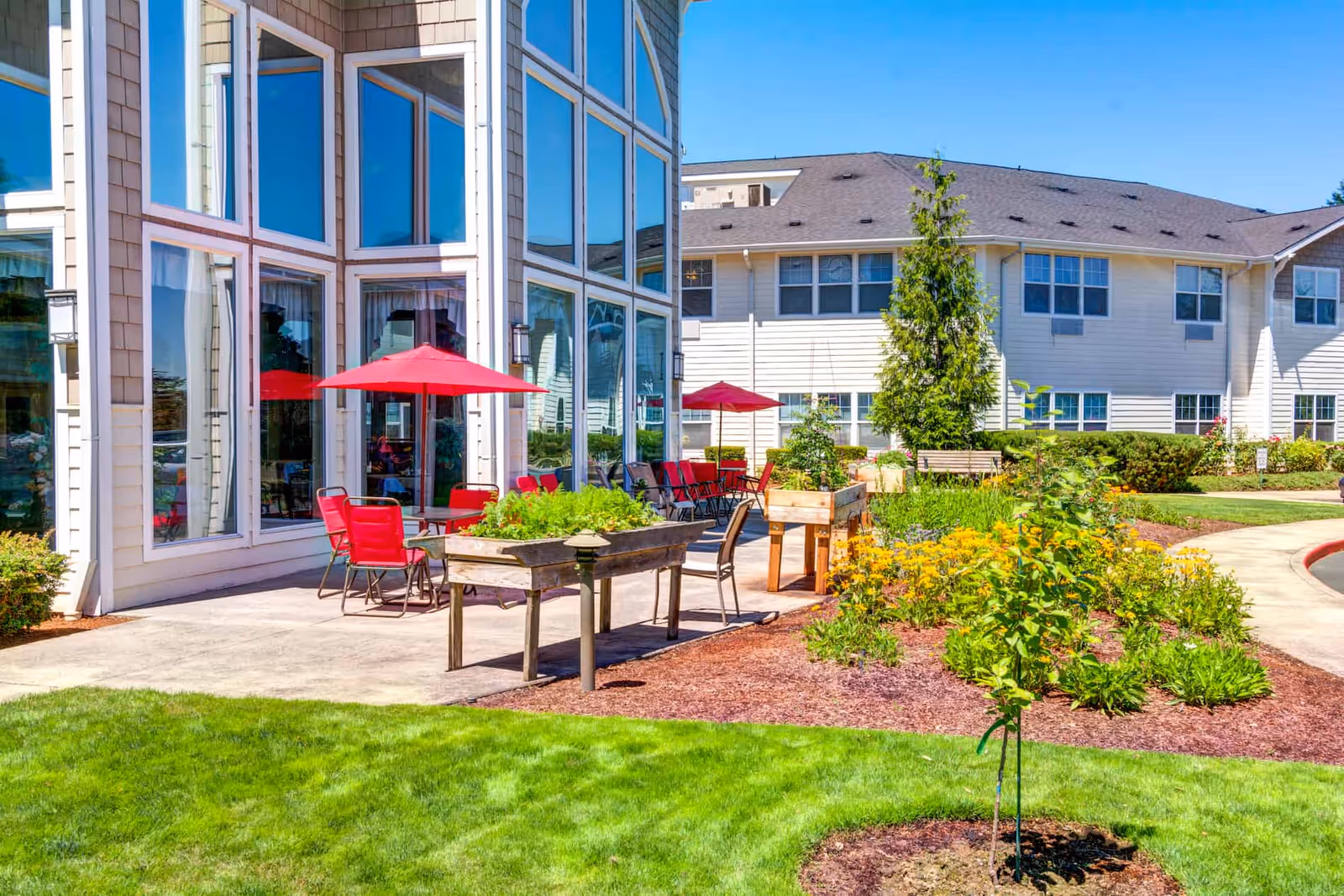 Outdoor patio and garden area with red umbrellas, seating, planter boxes, and large glass windows on a senior living building.