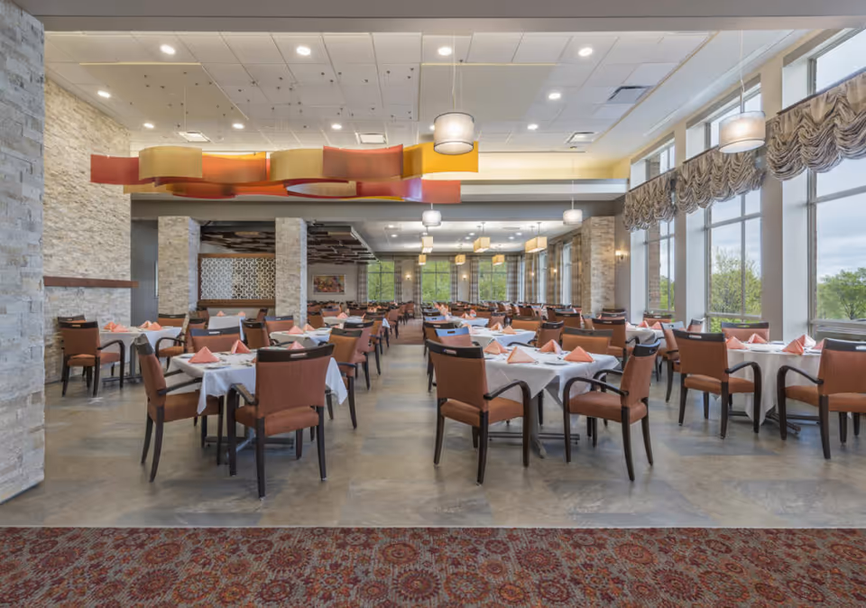Spacious dining room with multiple tables covered in white tablecloths and set with folded pink napkins. The room features large windows with draped curtains allowing natural light to fill the space, stone accent walls, and modern hanging light fixtures. Chairs with wooden frames and cushioned seats are arranged neatly around the tables.