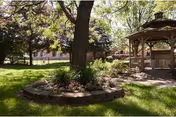 Sunlit courtyard with a large tree, a circular raised flower bed, a gazebo, and a grassy lawn in front of the facility building.