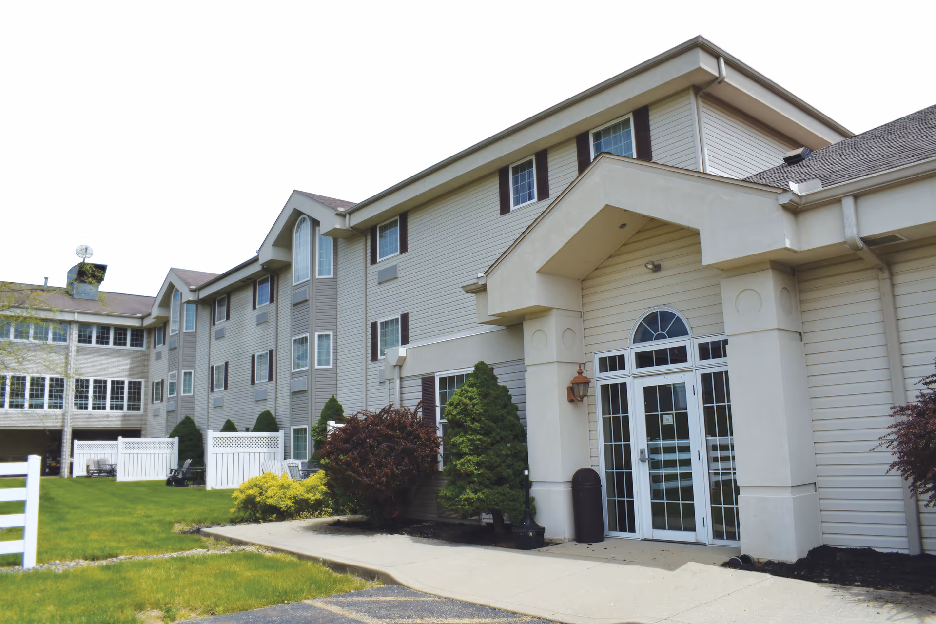 Exterior view of a multi-story senior living facility building with beige siding, multiple windows, and a main entrance featuring glass double doors under a covered portico. There are shrubs and small trees planted along the building, a grassy lawn, and a paved walkway leading to the entrance.