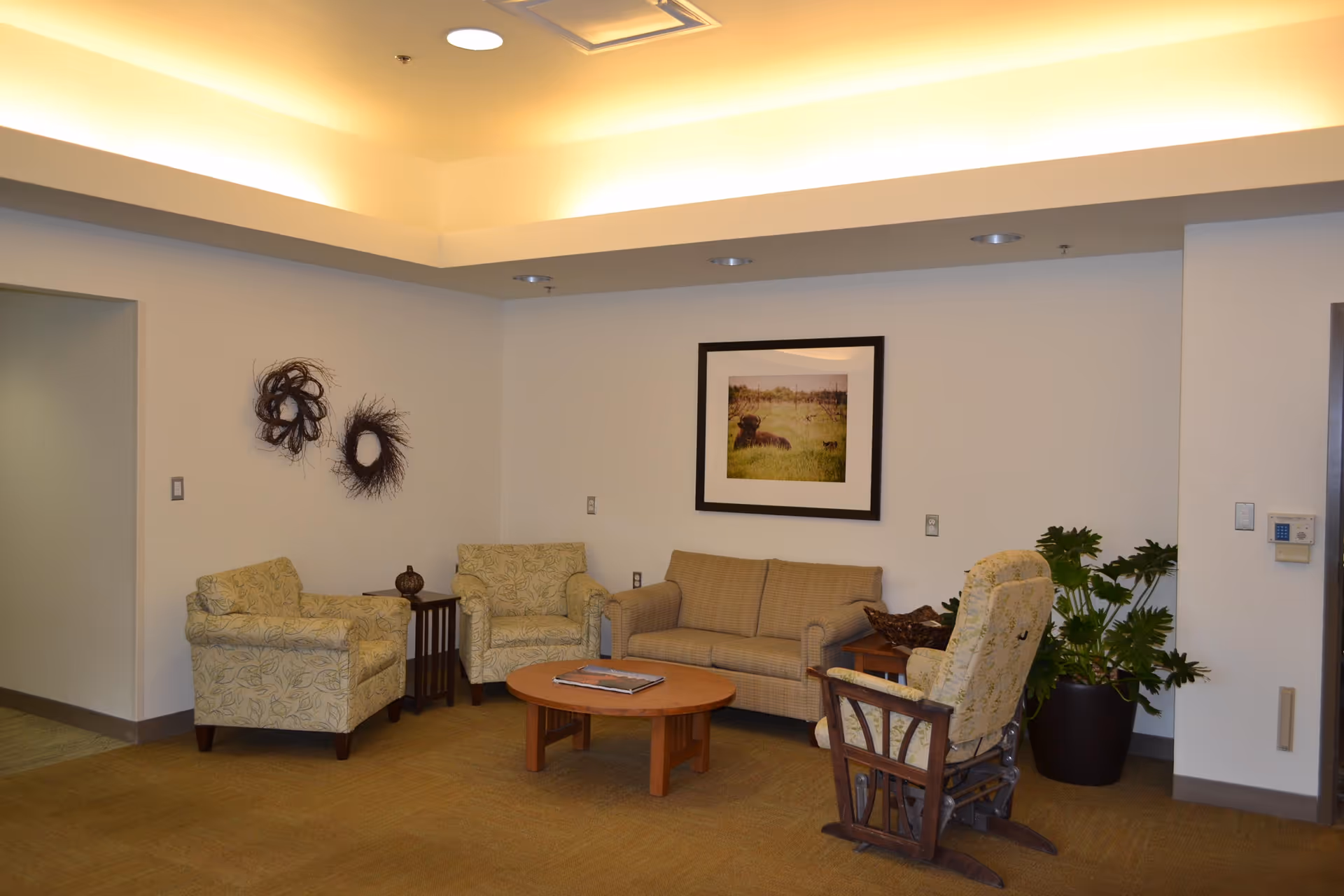 A cozy seating area in a senior living facility with two patterned armchairs, a beige sofa, a wooden coffee table with a book on it, a rocking chair, a side table with a decorative item, a large potted plant, and wall decorations including a framed picture of a dog in a field and two circular twig wreaths.