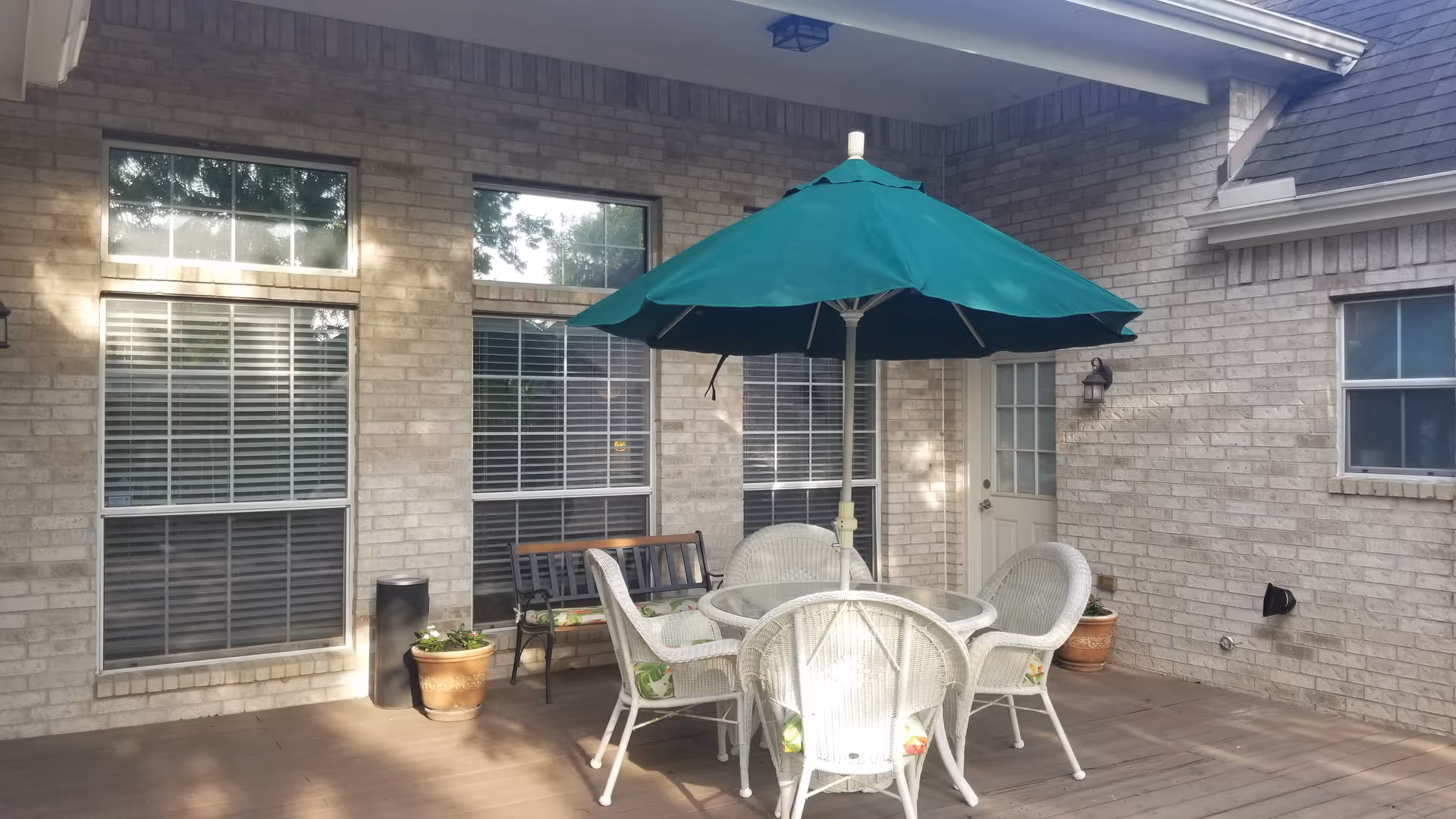 Outdoor patio area with a round glass table surrounded by four white wicker chairs with cushions. A large green umbrella is positioned over the table. There is a wooden bench against the brick wall of the building, two large windows with blinds, a door with a window, and potted plants on the patio.