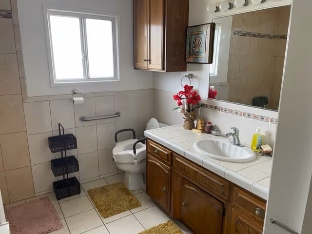 A bathroom with a white toilet equipped with safety rails, a white sink set in a tiled countertop with wooden cabinets below, a large mirror above the sink, a window letting in natural light, and various bathroom accessories including a pink flower arrangement, soap dispenser, and bath mats on the tiled floor.