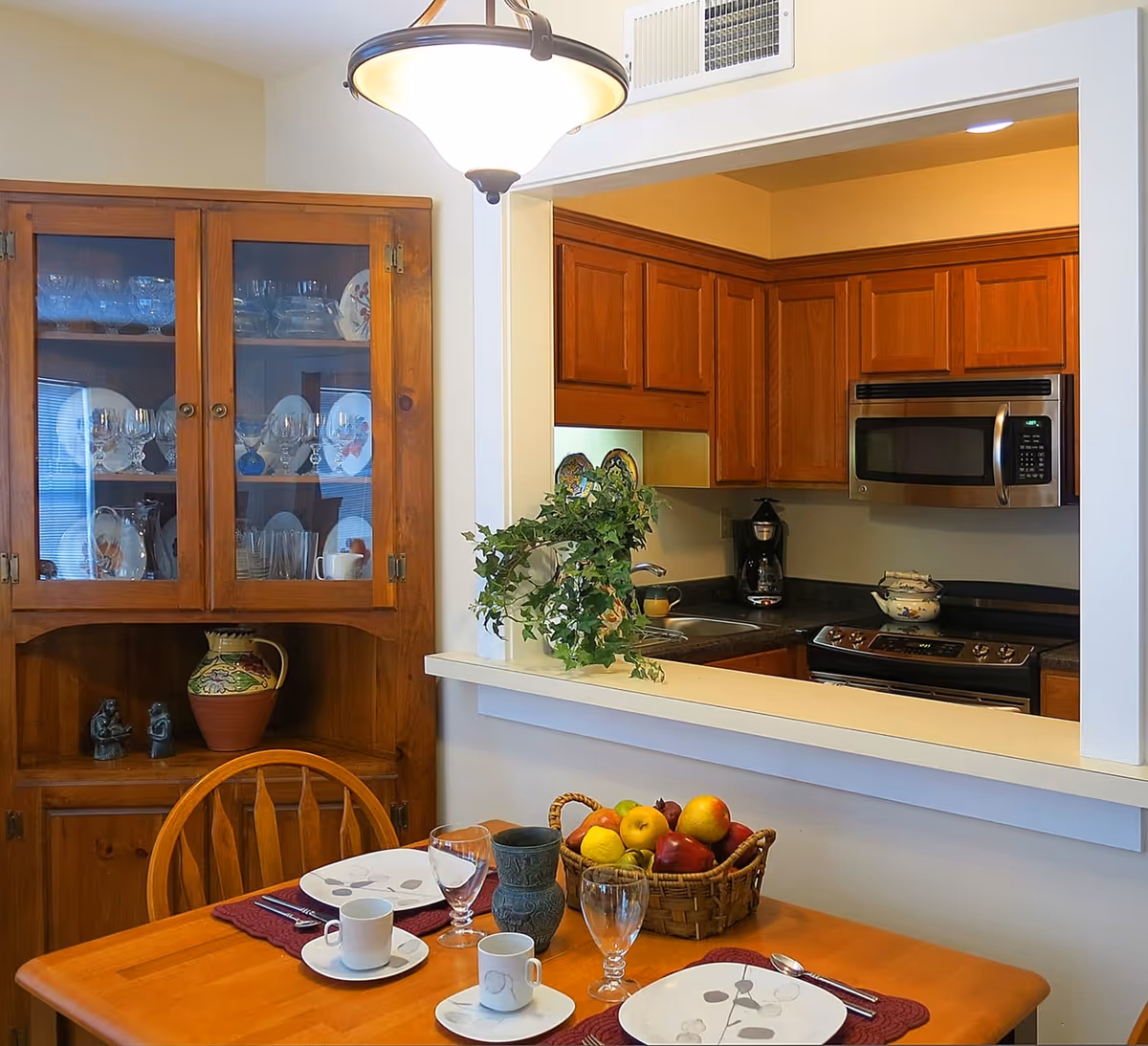 A cozy dining area with a wooden table set for two, featuring plates, cups, glasses, and cutlery. A basket of fresh fruit is placed in the center of the table. Behind the table is a wooden cabinet displaying glassware and decorative plates. The dining area opens into a kitchen with wooden cabinets, a microwave, a coffee maker, and a stove. A hanging light fixture illuminates the space.