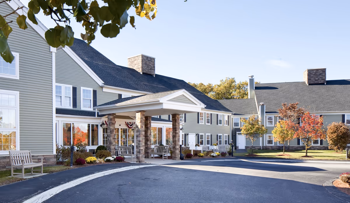Exterior view of a senior living facility building with a covered entrance supported by stone pillars. The building has light green siding, multiple windows, and a dark shingled roof. There are benches and landscaped bushes with flowers near the entrance, and trees with autumn foliage in the surrounding lawn area.