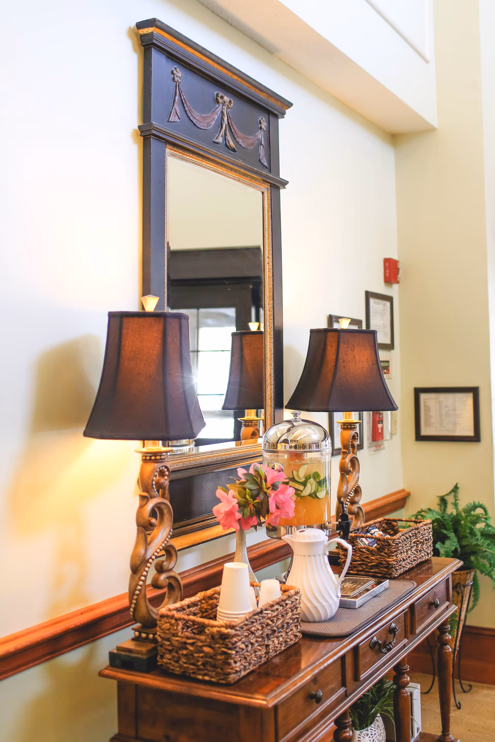 A wooden console table with two ornate lamps with black lampshades, a large decorative mirror above it, a glass beverage dispenser with fruit-infused water, a white pitcher, two woven baskets holding cups and other items, and a vase with pink flowers. The setting appears to be a well-lit interior space with framed documents on the wall and a potted plant nearby.
