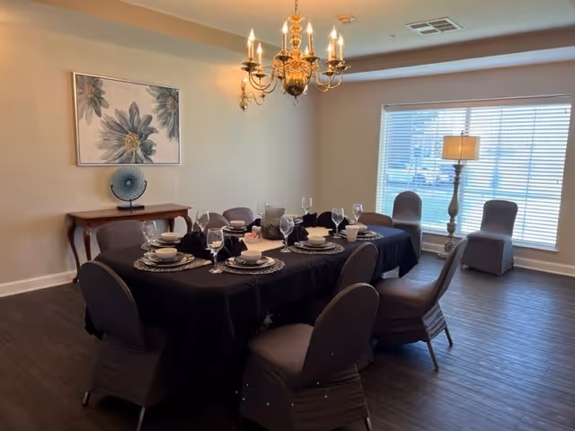 A dining room with a rectangular table covered with a black tablecloth set for eight people with plates, bowls, and wine glasses. The chairs around the table are covered with dark fabric. There is a large window with blinds letting in natural light, a floor lamp beside it, a wall-mounted chandelier above the table, and a small wooden console table against the wall with decorative items and a floral painting above it.