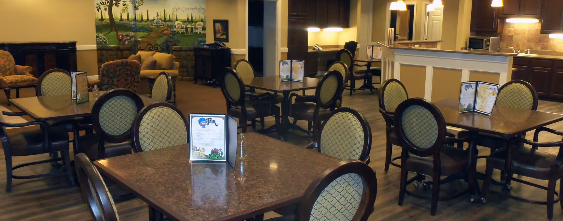 A dining area in a senior living facility with several tables and chairs arranged neatly. The tables have menus placed on them. In the background, there is a small kitchen area with cabinets, a microwave, and a sink. There is also a cozy seating area with armchairs and a mural on the wall depicting an outdoor garden scene.