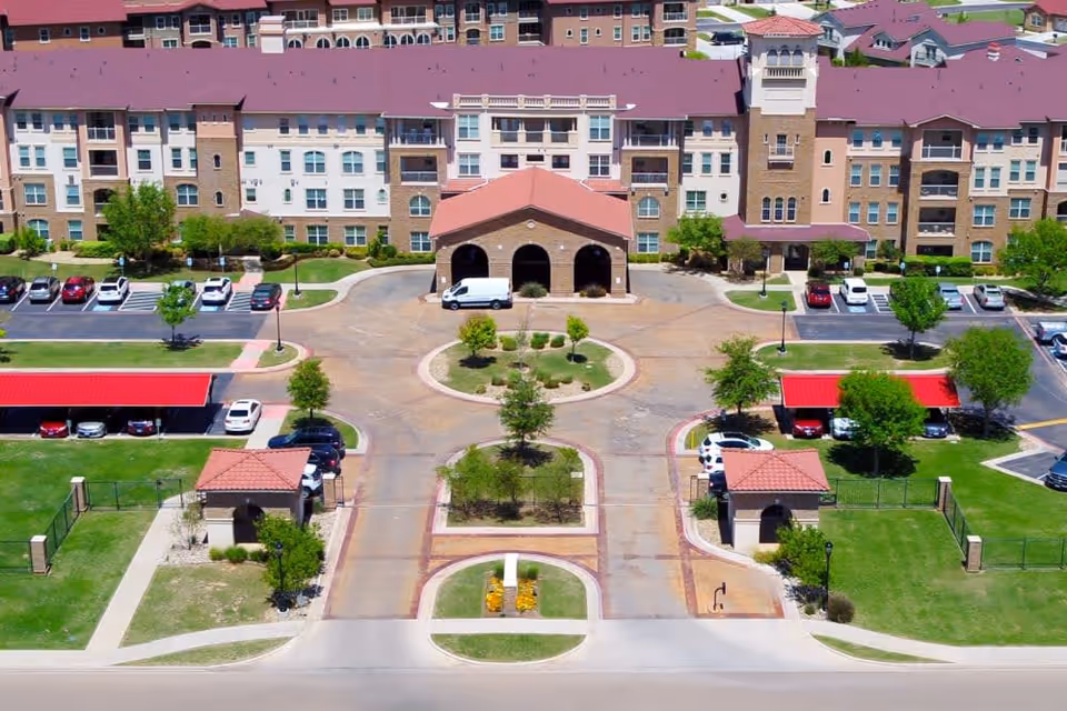 Aerial view of Isle at Raider Ranch senior living facility showing a large multi-story building with a red roof, a circular driveway with landscaped greenery in the center, parking lots with cars, covered parking areas, and small gated garden or courtyard areas in the front.