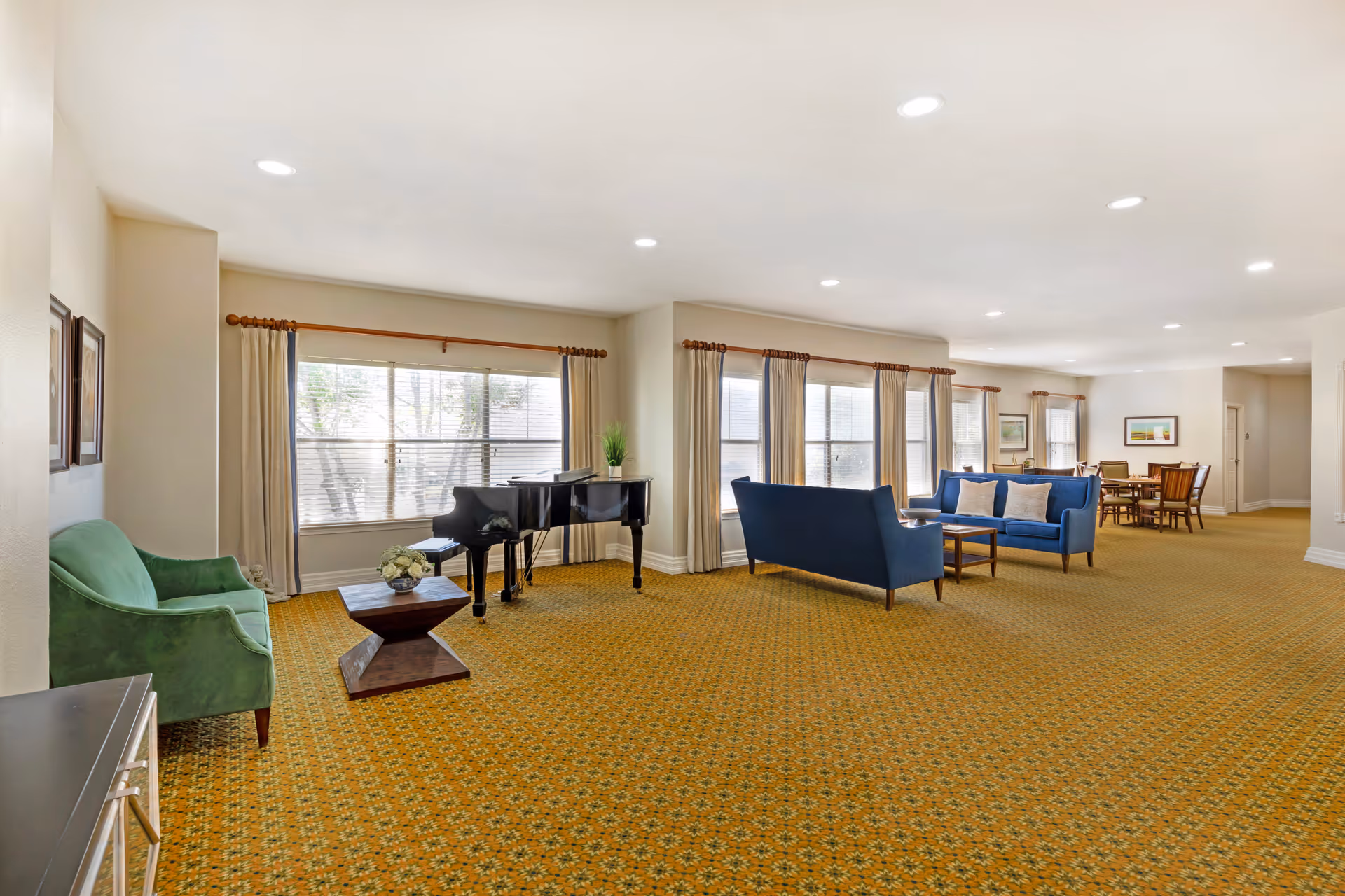 A spacious and well-lit common area with large windows covered by beige curtains. The room features a green armchair, a black grand piano, two blue sofas with white pillows, a wooden coffee table, and a dining table with chairs in the background. The floor is covered with a patterned yellow carpet, and the ceiling has recessed lighting.