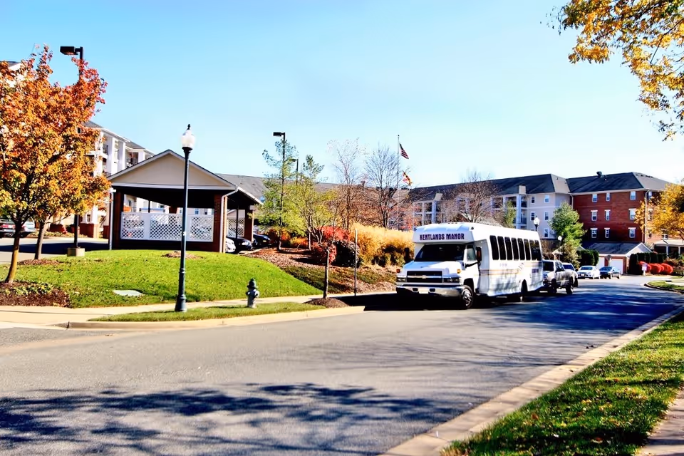 Exterior view of Kentlands Manor facility with a white shuttle bus parked on the road. The building is multi-story with a mix of brick and light-colored siding. Trees with autumn foliage and a small covered pavilion are visible on a grassy area beside the road under a clear blue sky.