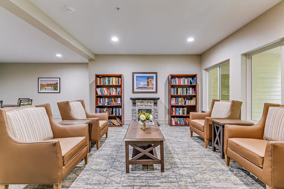 A cozy senior living common area with four tan armchairs arranged around a wooden coffee table. Behind the table is a stone fireplace flanked by two wooden bookshelves filled with books. The walls are light-colored, and there are large windows on the right side letting in natural light. A framed picture hangs above the fireplace.