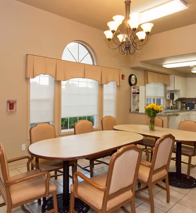 A dining area with two oval tables surrounded by cushioned wooden chairs. The room has beige walls, a large arched window with beige valances and white blinds, and a chandelier with multiple lights hanging from the ceiling. In the background, there is a kitchen area with white cabinets and a countertop, and a vase with yellow flowers on one of the tables.