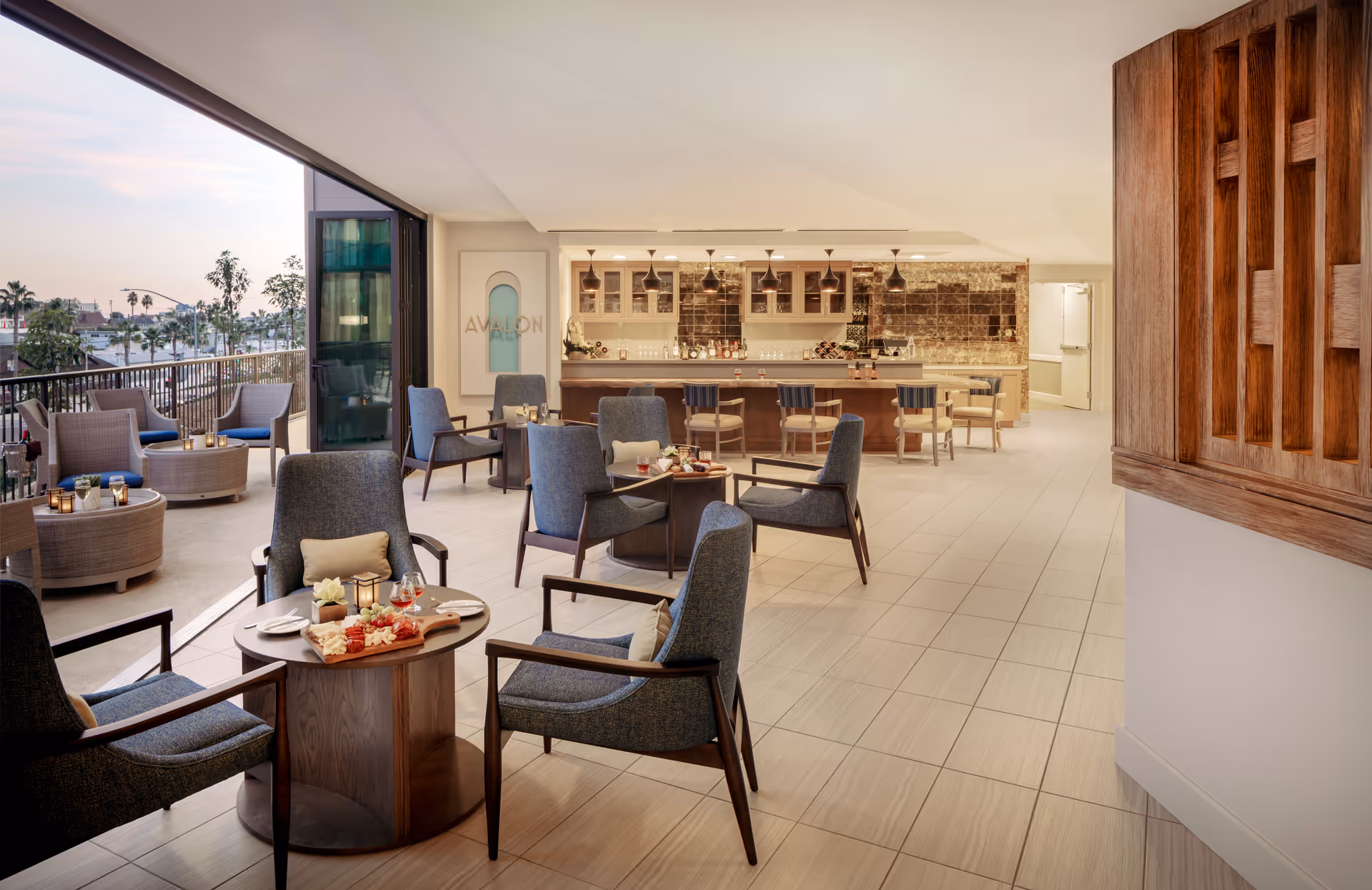 A modern lounge area with several blue cushioned armchairs and small round tables set with snacks and drinks. The space opens to an outdoor patio with additional seating and a view of palm trees and buildings. In the background, there is a bar with stools and pendant lighting, and a sign on the wall that reads 'Avalon'.