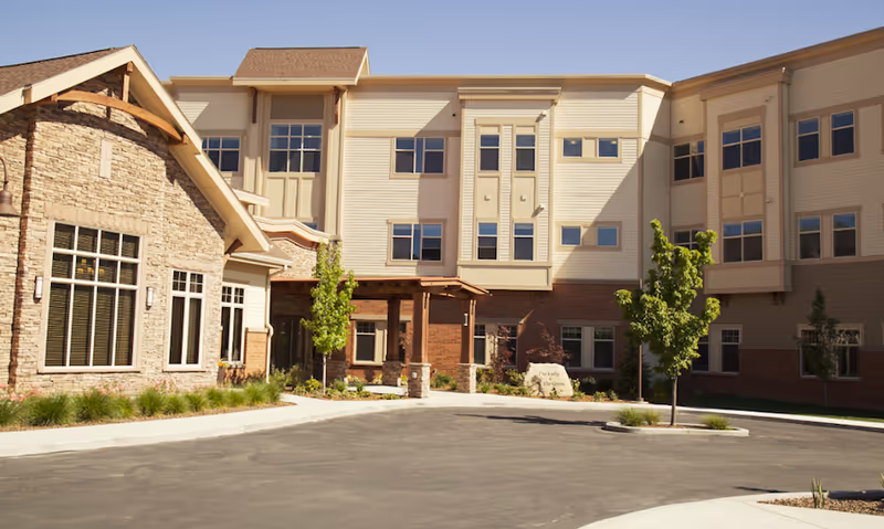Exterior view of a senior living facility building with beige siding and stone accents, multiple windows, a covered entrance, small trees, and landscaping under a clear blue sky.