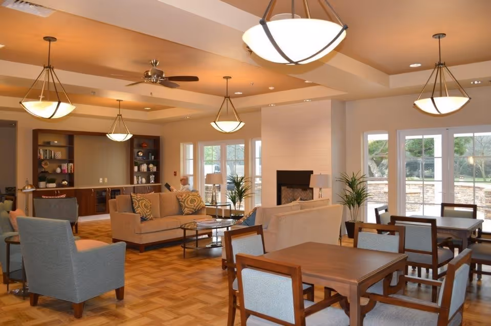 A spacious and well-lit common area in a senior living facility featuring comfortable beige sofas, gray armchairs, wooden tables with chairs, a fireplace, large windows and glass doors overlooking an outdoor garden, and ceiling lights with fans.