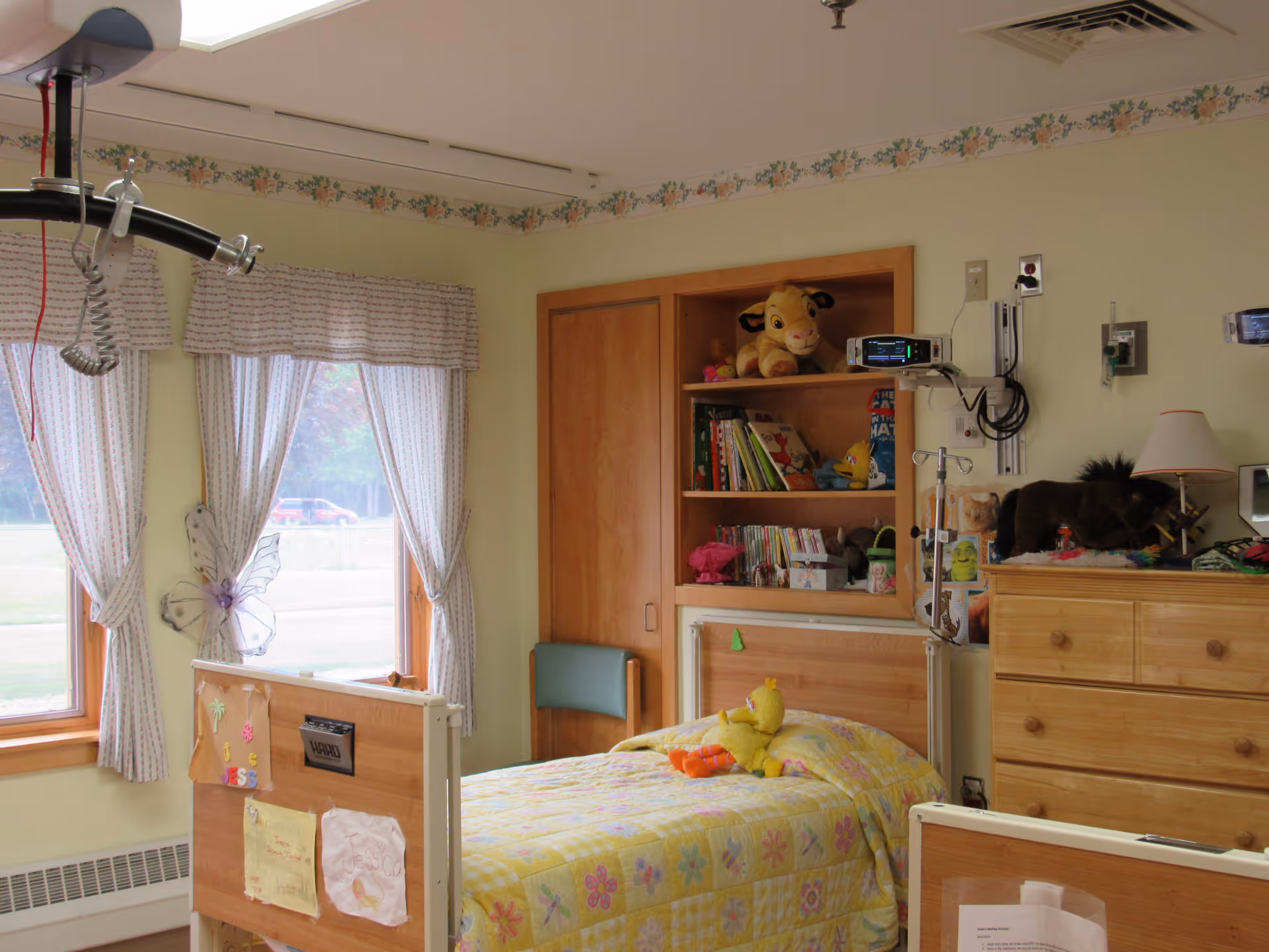 A bright, homey patient bedroom with a single hospital-style bed covered in a floral bedspread, stuffed animals, a bookshelf and wooden dresser.