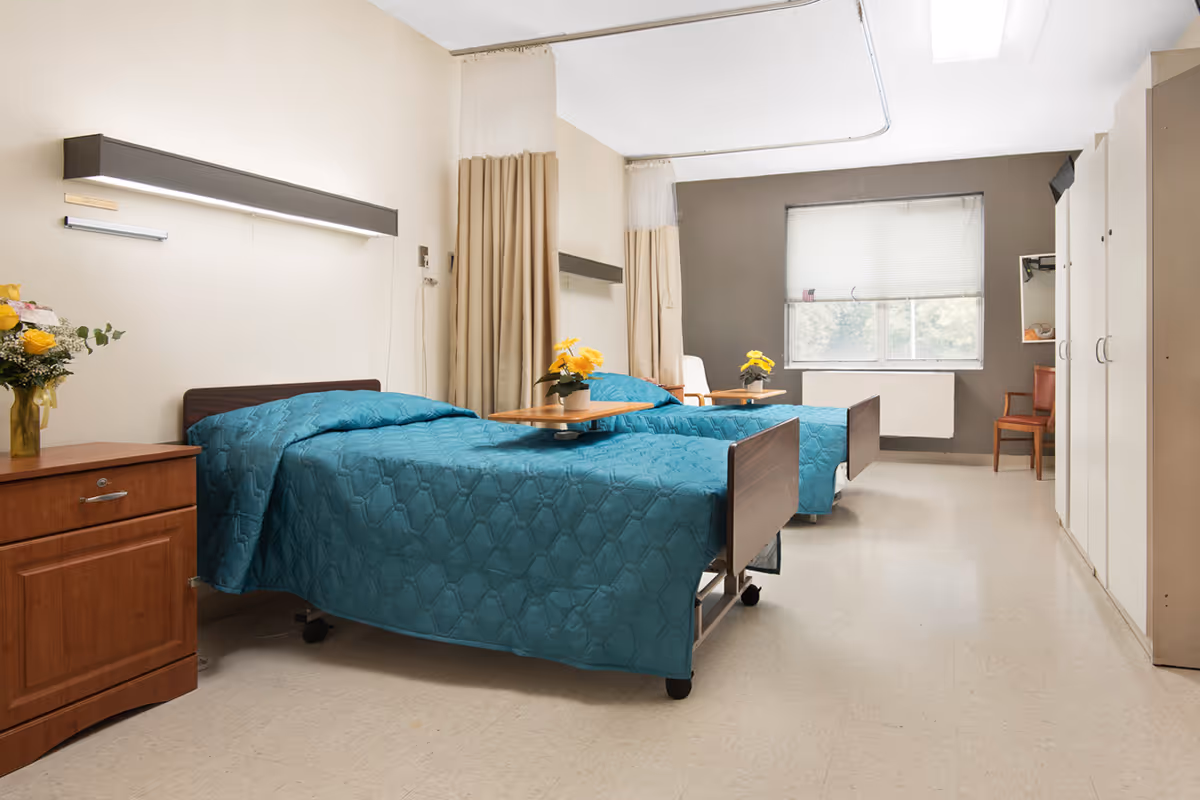 A bright and clean nursing facility room with two hospital-style beds covered in teal quilts. Each bed has a small wooden overbed table with a yellow flower pot on top. There is a wooden nightstand with a vase of flowers on the left side, beige curtains between the beds, a window with blinds at the far end, and white wardrobes along the right wall. The room has a neutral color palette with beige and brown tones.