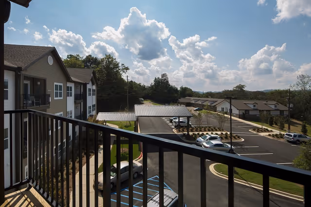 View from a balcony overlooking a parking lot with several parked cars and covered parking spaces. The scene includes a multi-story residential building on the left and single-story buildings in the background under a partly cloudy sky.
