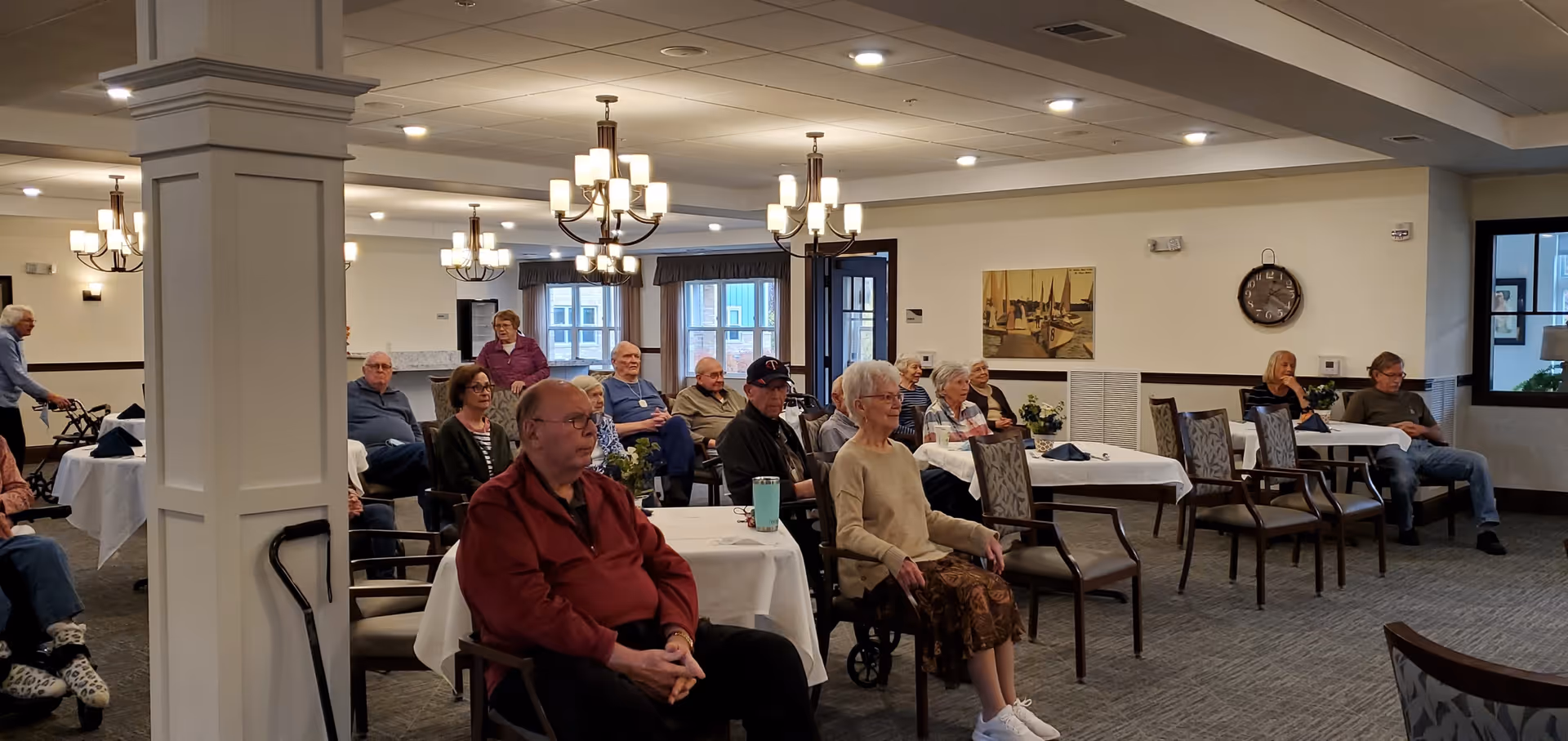 A group of elderly people seated in a well-lit common area with tables covered in white tablecloths and chairs arranged facing forward. The room has chandeliers hanging from the ceiling, a large pillar in the middle, and windows letting in natural light. Some people are sitting in wheelchairs or using walkers.