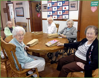 Five elderly women sit around a round wooden table in a communal room with paper bags and a quilted wall hanging behind them.