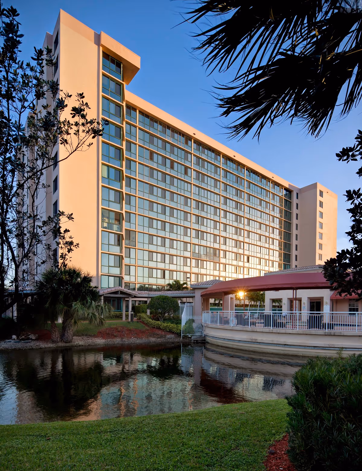 A tall sunlit multi-story building reflected in a pond with landscaped grounds and a covered terrace.