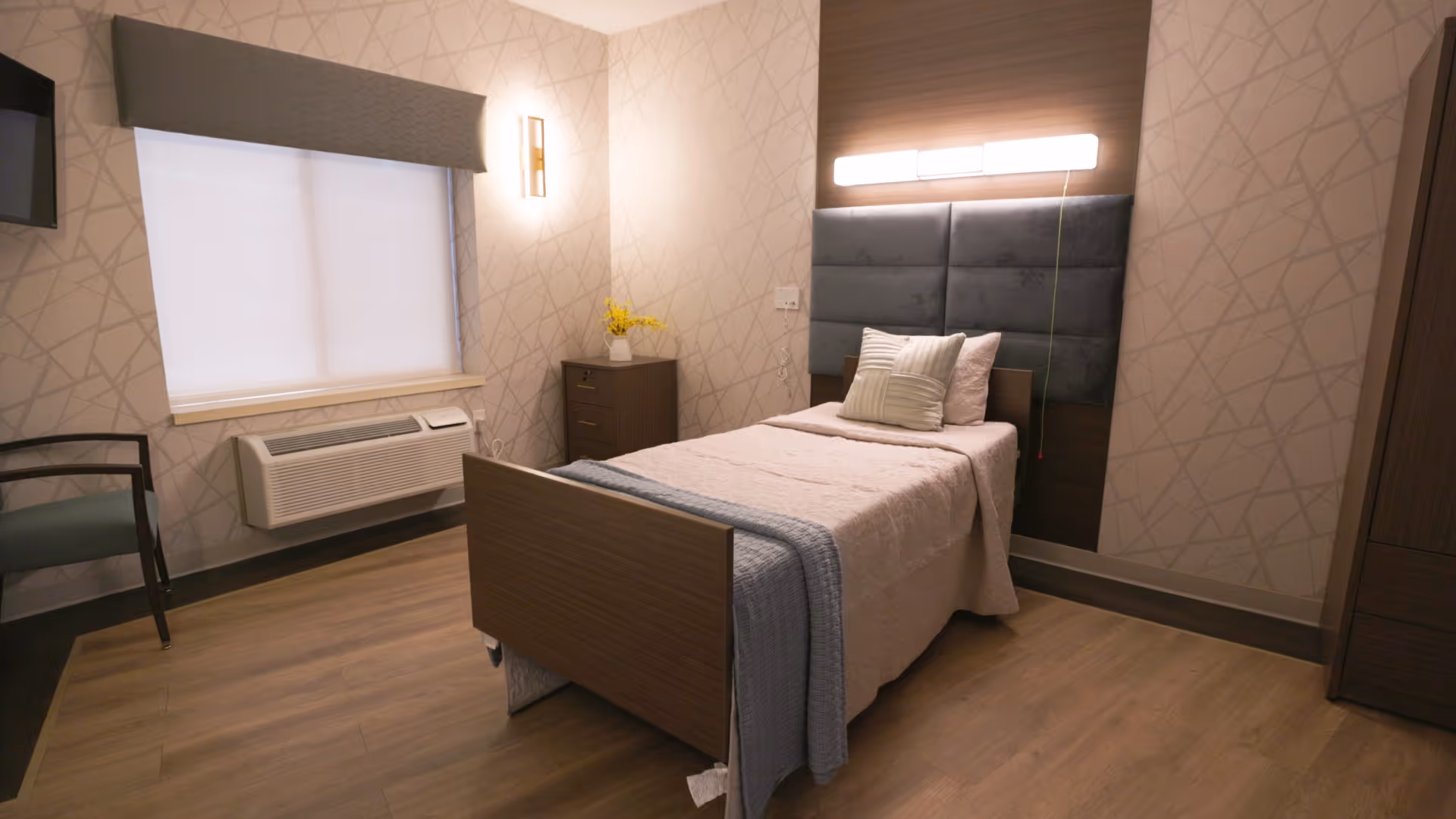 A neatly arranged single bed in a senior living facility room with a padded headboard, a bedside cabinet with a vase of yellow flowers, a chair, a window with a blind, and a wall-mounted air conditioning unit.