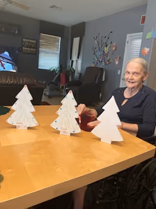 An elderly woman sitting at a wooden table in a living area, smiling and holding a paper Christmas tree decoration. Three paper Christmas trees are placed on the table in front of her. The room has gray walls, a window with blinds, a dark armchair, and wall decorations including a tree with colorful leaves.