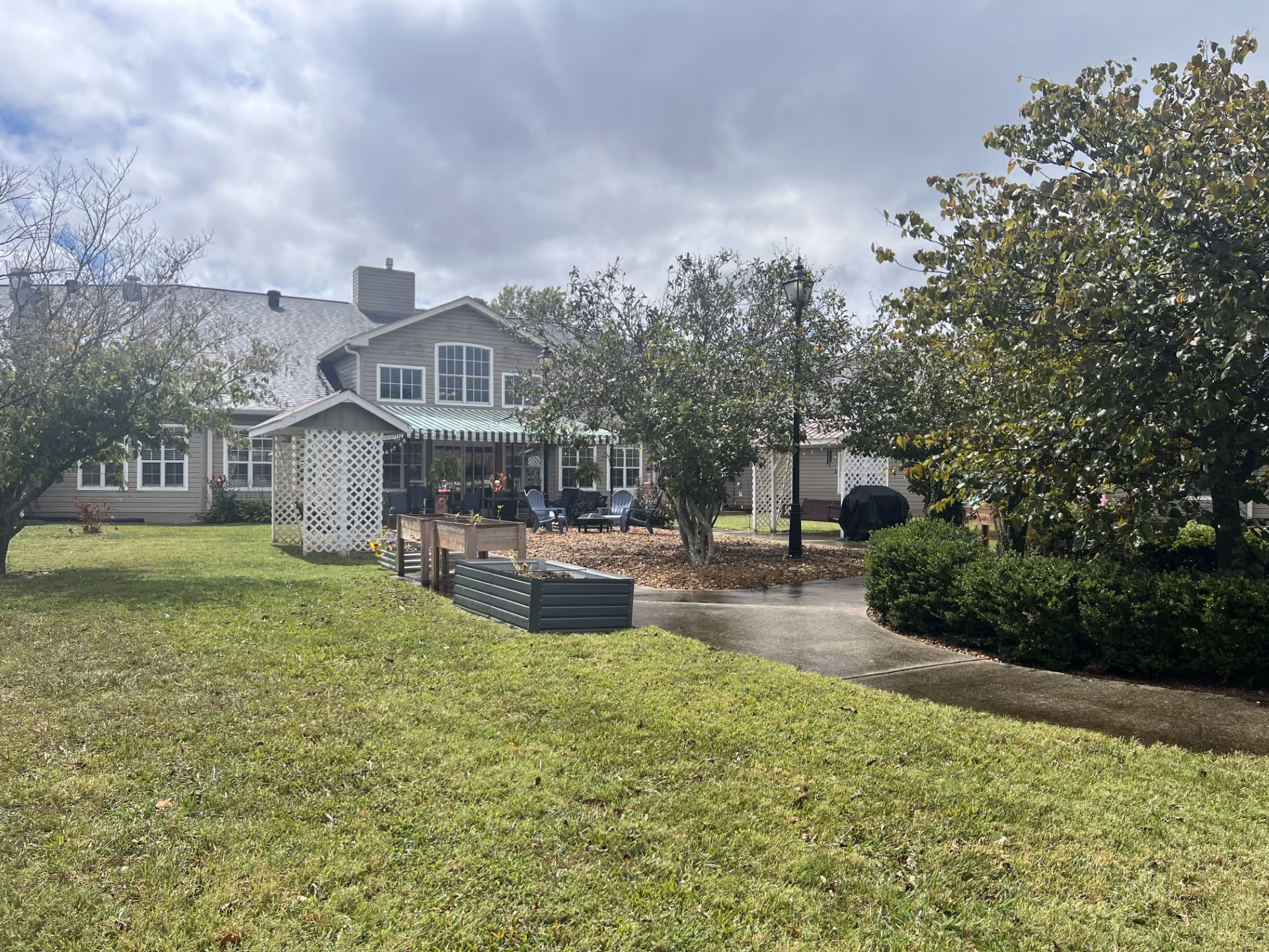 Grassy courtyard with raised garden beds, trees, a seating area and a two-story building in the background under a cloudy sky.