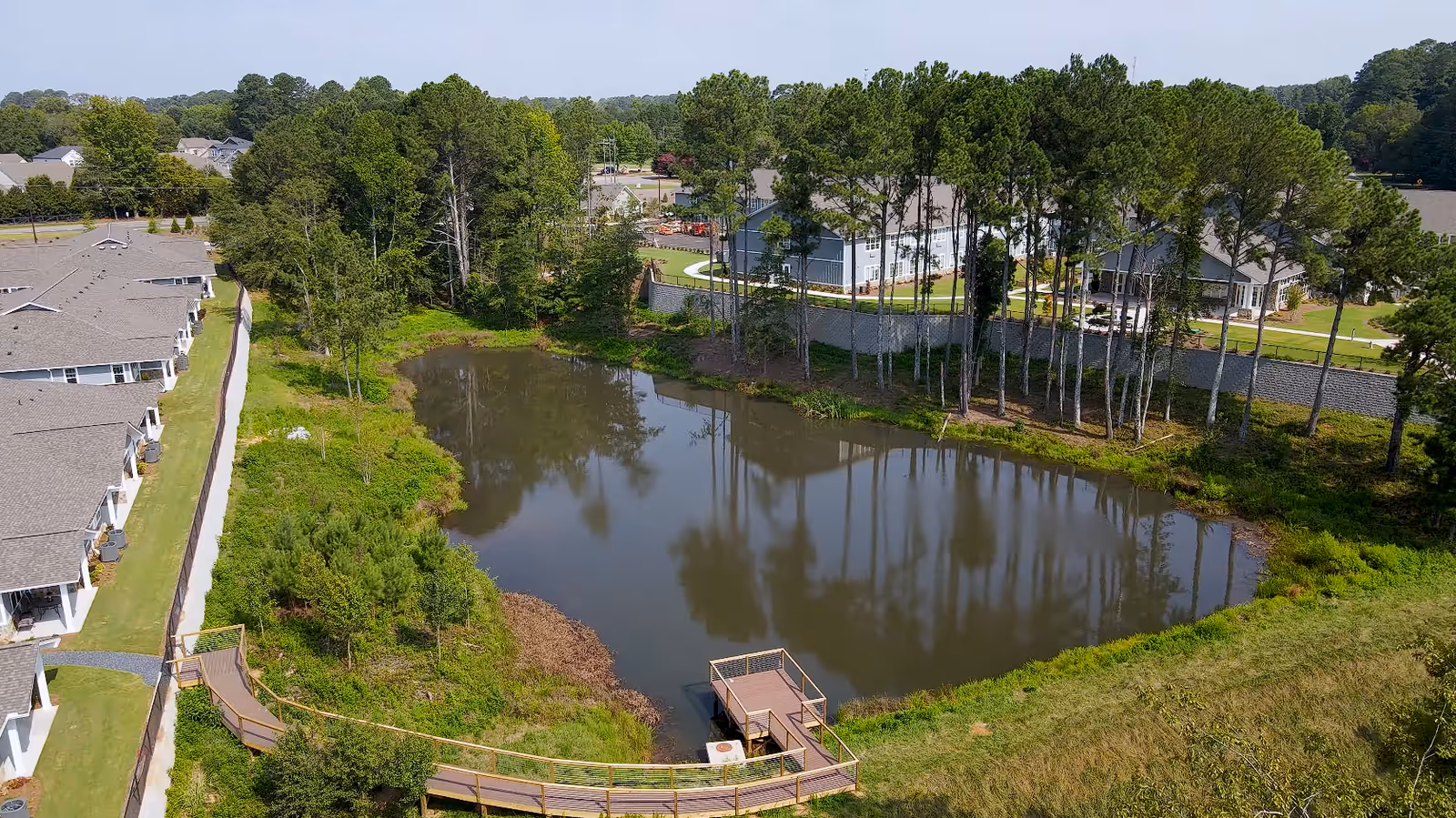 Aerial view of a pond surrounded by trees and greenery with a wooden dock extending into the water. Residential buildings and a facility are visible in the background behind a retaining wall and a row of tall pine trees.