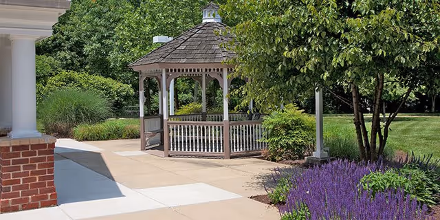 A wooden gazebo on a paved courtyard surrounded by shrubs, trees, and purple flowering plants.